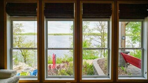 A room with a large window overlooking a lake, a red chair, and a small table. - Morning Sun Cabin on Lake Metigoshe (Bottineau)