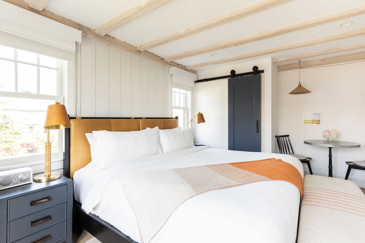 A neatly made bed with white linens, a bedside table with a lamp, and a small table with a vase of flowers.