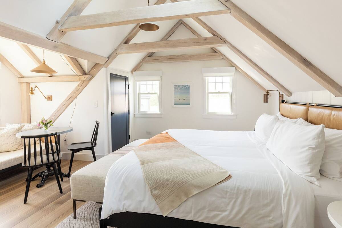 A bedroom with a large bed, a small table with chairs, and a view of the outside through a window.