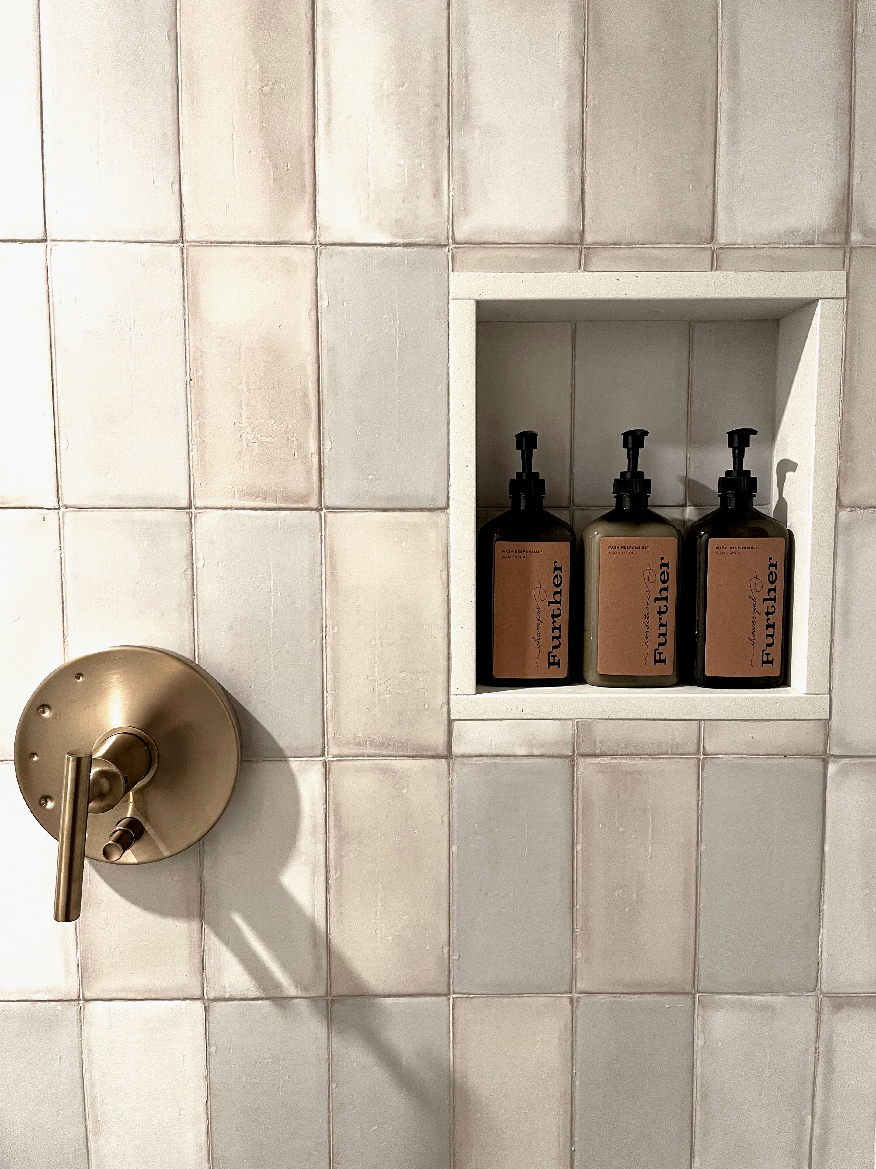 Un baño con pared de azulejos blancos, un grifo dorado y tres botellas de jabón para manos sobre un estante blanco.