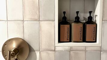 Un baño con pared de azulejos blancos, un grifo dorado y tres botellas de jabón para manos sobre un estante blanco.