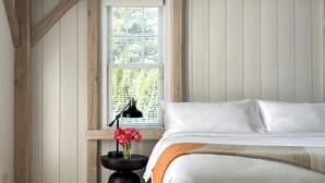 Een slaapkamer met een houten plafond, een groot raam, een nachtkastje met een lamp en een bed met wit beddengoed.