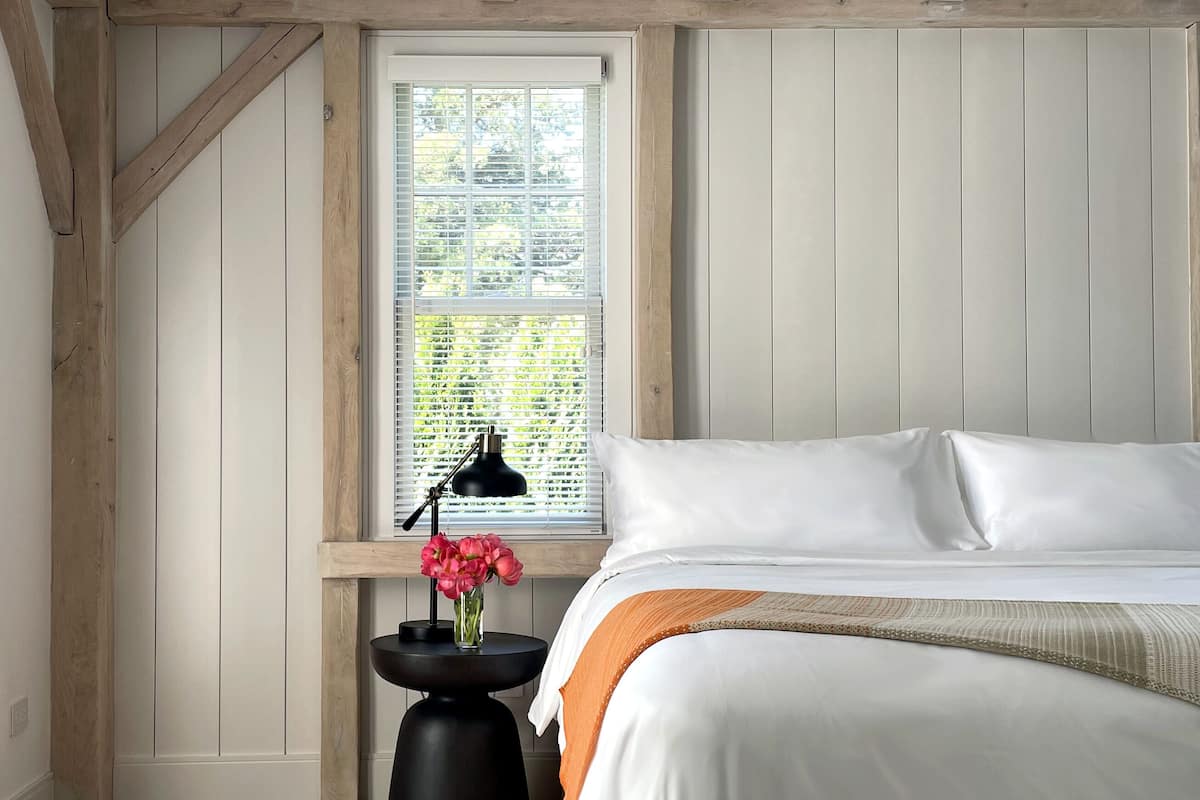 A bedroom with a wooden ceiling, a large window, a bedside table with a lamp, and a bed with white bedding.