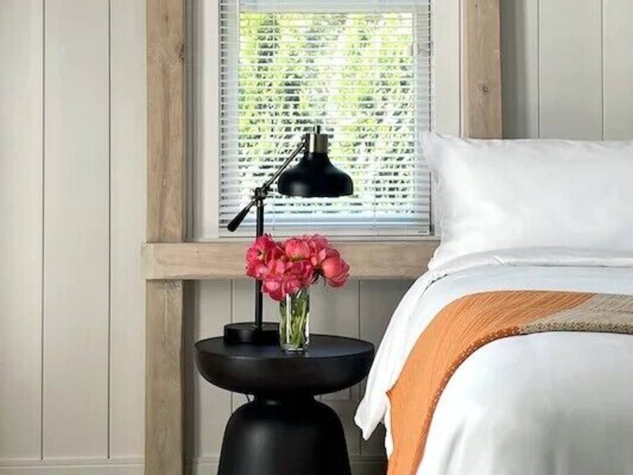 A neatly made bed with white linens and a beige throw blanket, a black lamp on a small table beside the bed, and a vase with pink flowers on the table.