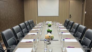 Un salón de conferencias con una larga mesa, sillas de cuero negro, botellas de agua y una pantalla para proyector.