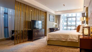 A modern hotel room with a large bed, a television, a desk, and a view of the city through large windows.