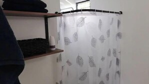 Une salle de bain avec un rideau de douche orné d’un motif de feuilles, une étagère avec des vêtements pliés et une bouteille posée sur l’étagère.