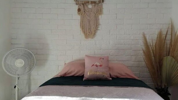 Une chambre avec un mur en briques blanches, une décoration murale en macramé, un lit avec une literie rose et verte, et un ventilateur sur pied.