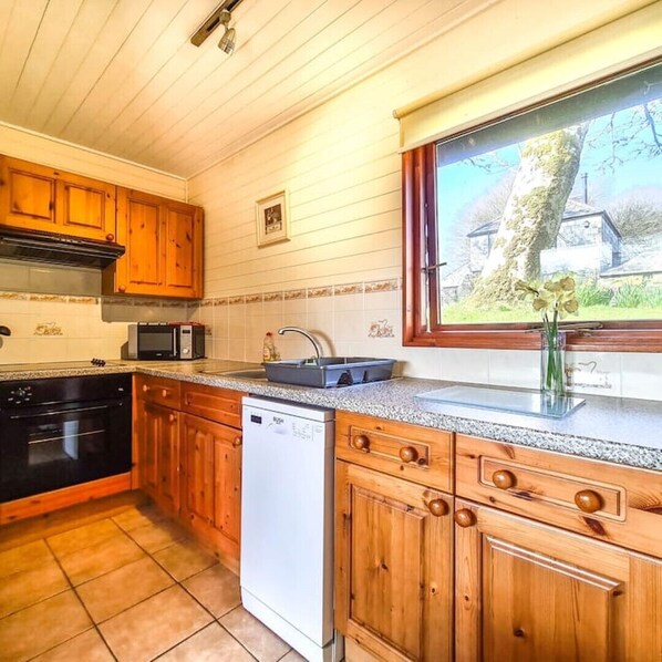 A kitchen with wooden cabinets, a microwave, a dishwasher, and a window with a view of a tree and a house. - Ta Mill Brookview Lodge 2 (Launceston)