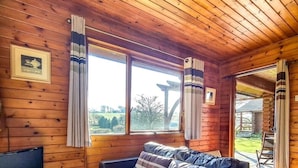 A cozy cabin interior with a leather sofa, wooden walls, a television, and large windows offering a view of the outdoors. - Ta Mill Brookview Lodge 2 (Launceston)