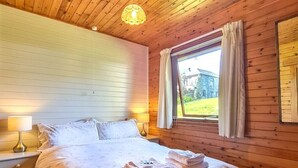 A wooden cabin bedroom with a bed, two bedside lamps, a window with curtains, and a view of a building outside. - Ta Mill Brookview Lodge 2 (Launceston)