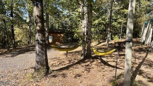 Un hamac est suspendu entre deux arbres dans une zone boisée, avec un petit bâtiment visible au loin.