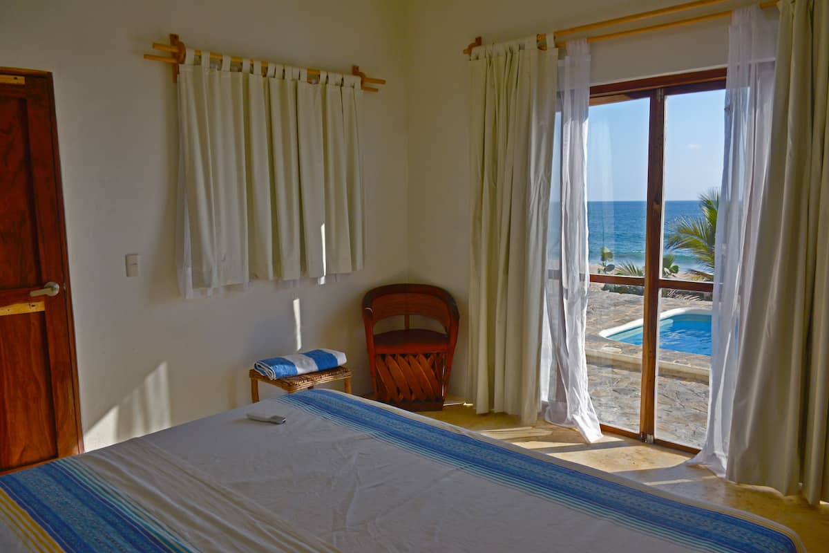 Une chambre à coucher comprenant un lit, une chaise, une petite table et une vue sur l’océan, visible à travers une porte-fenêtre coulissante.