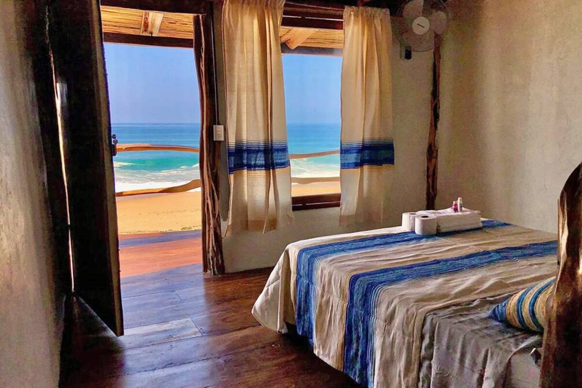 Une chambre avec un lit, un plancher en bois et une vue sur la plage, visible à travers les rideaux.
