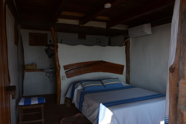Un dormitorio con cabecera de madera, cama con ropa de cama azul y blanca, un taburete de madera pequeño, un lavamanos y un aire acondicionado instalado en la pared.