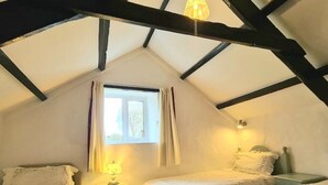 A cozy attic bedroom with two beds, a window with curtains, a lamp, and a hanging light fixture. - Ta Mill Forge Cottage in a Stunning Retreat (Launceston)
