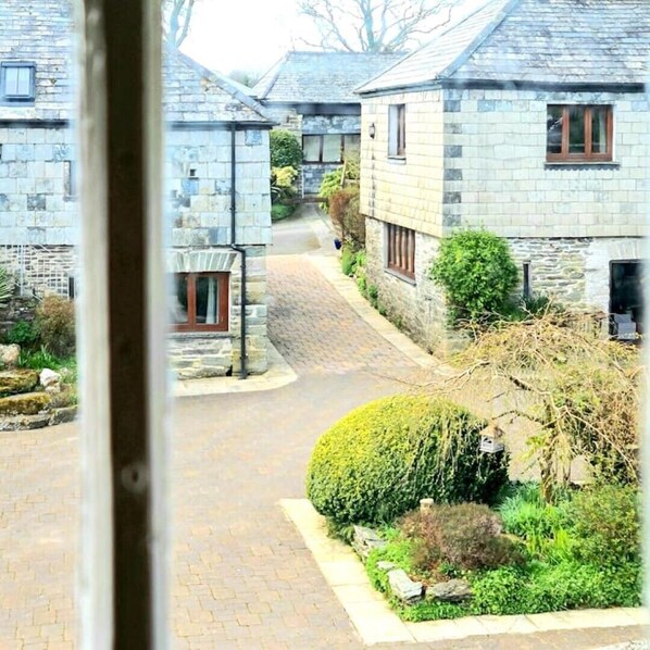 Un bâtiment en pierre donnant sur une rue pavée, un petit jardin avec un arbuste bien taillé et une fenêtre visible avec un cadre en bois.