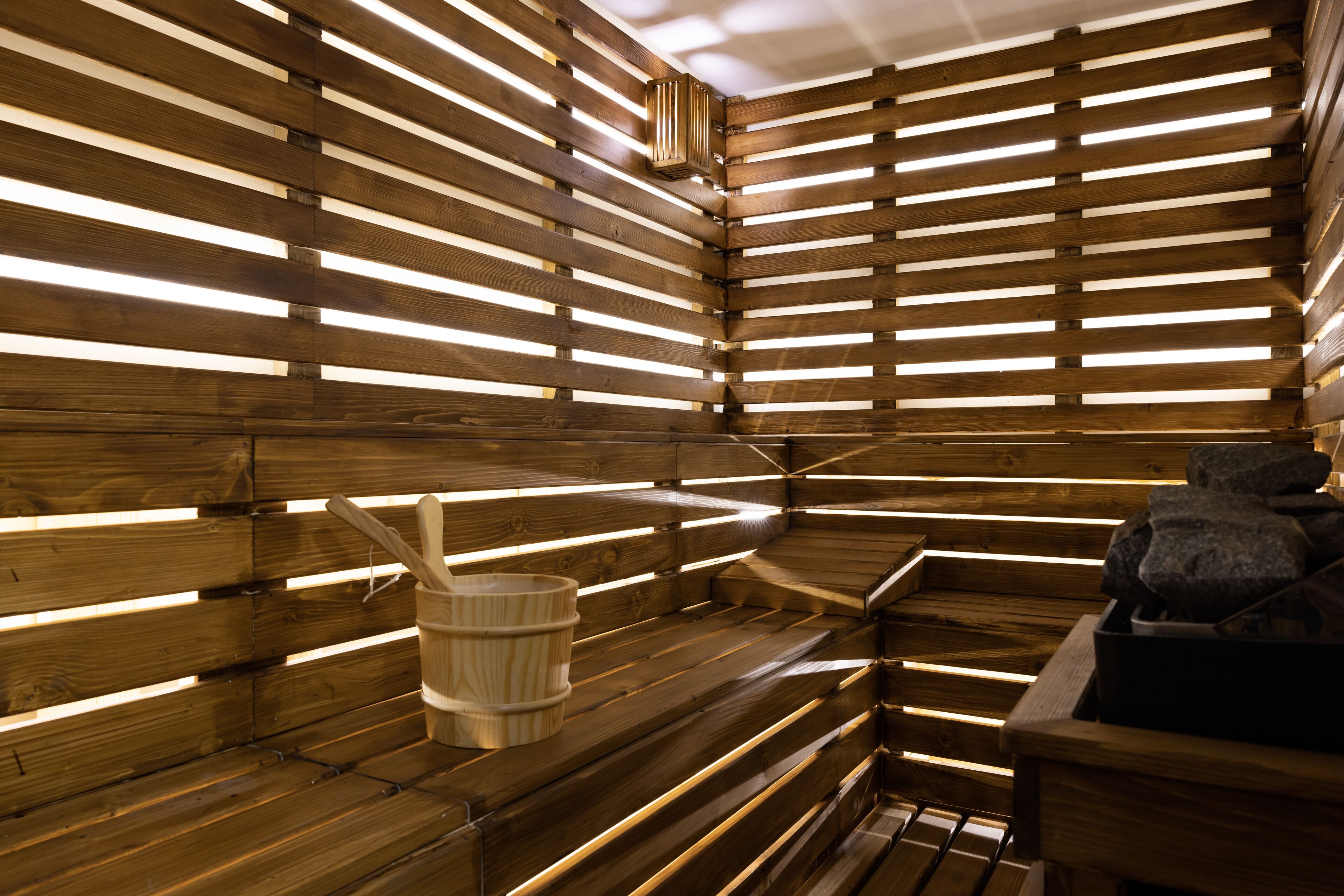A sauna with wooden slats and benches, a bucket, and a stone container.