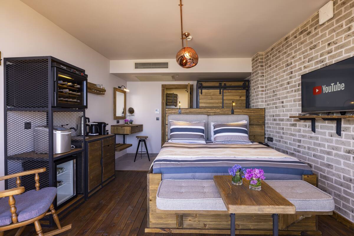 Une chambre moderne comprenant un lit, une table en bois, une chaise, un téléviseur et un coin cuisine avec un réfrigérateur et des étagères.