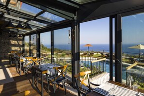 A dining area with tables and chairs, large windows, and a view of the sea. - Hotel Vita Naturalis (Ayvacik)