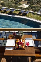 Una mesa con una bandeja de frutas, copas de vino y una planta en maceta en un balcón con vistas a una piscina y sillones de descanso.