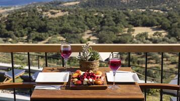 A balcony with a wooden table set for a meal, featuring a fruit platter and two glasses of red wine, overlooking a scenic landscape with hills and trees.