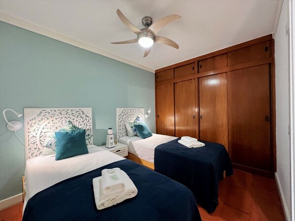 Habitació d'hotel amb dos llits, ventilador de sostre, armari de fusta i una petita taula de nit.