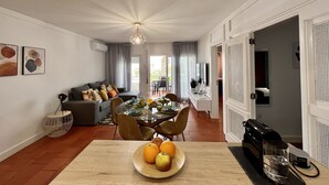 A modern living room with a dining area, featuring a wooden table with fruit, a sofa with cushions, and a coffee machine.