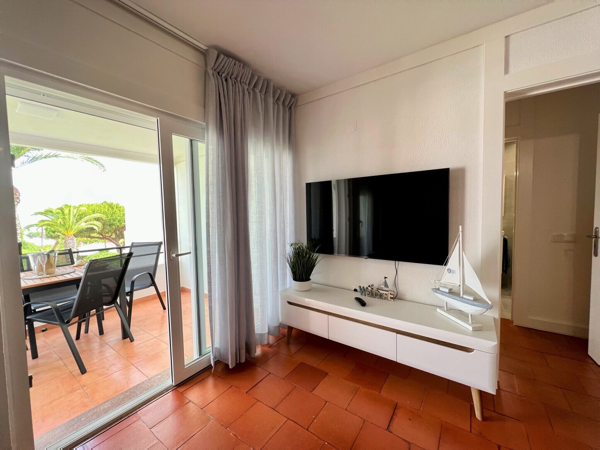 Una sala de estar moderna con un televisor de pantalla plana grande montado en la pared, un ventanal corredizo que da a un área exterior y un mueble de entretenimiento blanco con una planta pequeña y objetos decorativos.