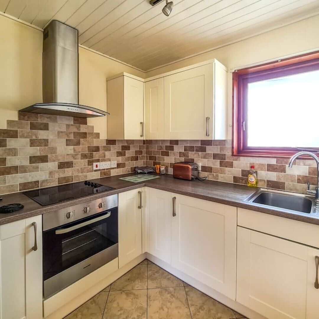 Una cocina con gabinetes blancos, un extractor de acero inoxidable, un horno integrado, una encimera con un tostador y un ventanal con marco rojo.