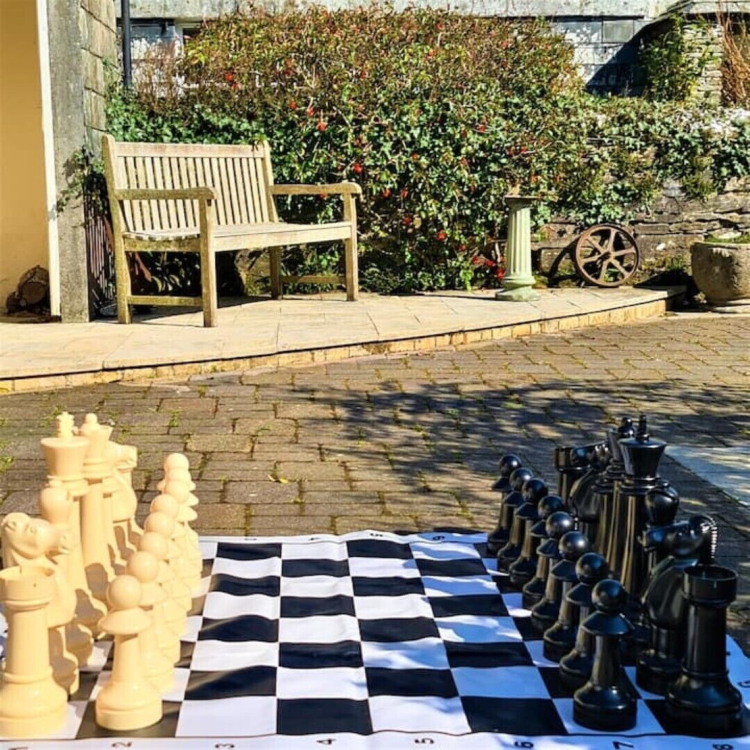 Ett schackbräde placerat utomhus på en kullerstensbelagd yta, med en bänk och grönska i bakgrunden.