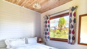 A bedroom with a large bed, a window with patterned curtains, and a view of greenery outside. - Cosy log-cabin style lodge with outdoor Lay-Z-Spa in rural, North Cornwall. (Launceston)