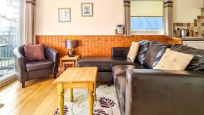 A cozy living room with a leather sofa, a wooden coffee table, and a small side table. There are two framed pictures on the wall, a window with curtains, and a view of the outside. - Ta Mill Holidays - Meadowview Chalet 3 (Launceston)
