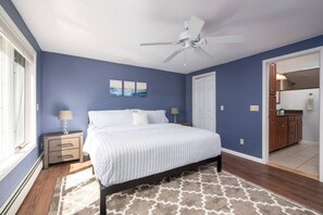 Un dormitorio con cama, mesitas de noche, ventilador de techo y vista a la cocina.