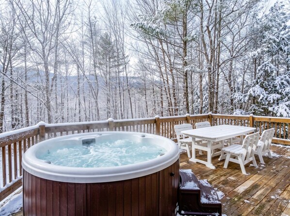 A hot tub on a wooden deck with a table and chairs, surrounded by snow-covered trees.