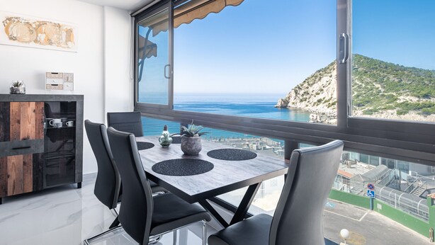 A modern dining area with a table and chairs, a wooden cabinet, and a large window offering a view of the sea and a hilly landscape.