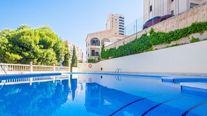 A swimming pool with clear blue water, surrounded by a white wall and a wooden fence. There are trees and buildings in the background.