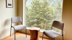 Una habitación con dos sillas grises, una mesa de centro de madera y un ventanal con vistas a una montaña boscosa.