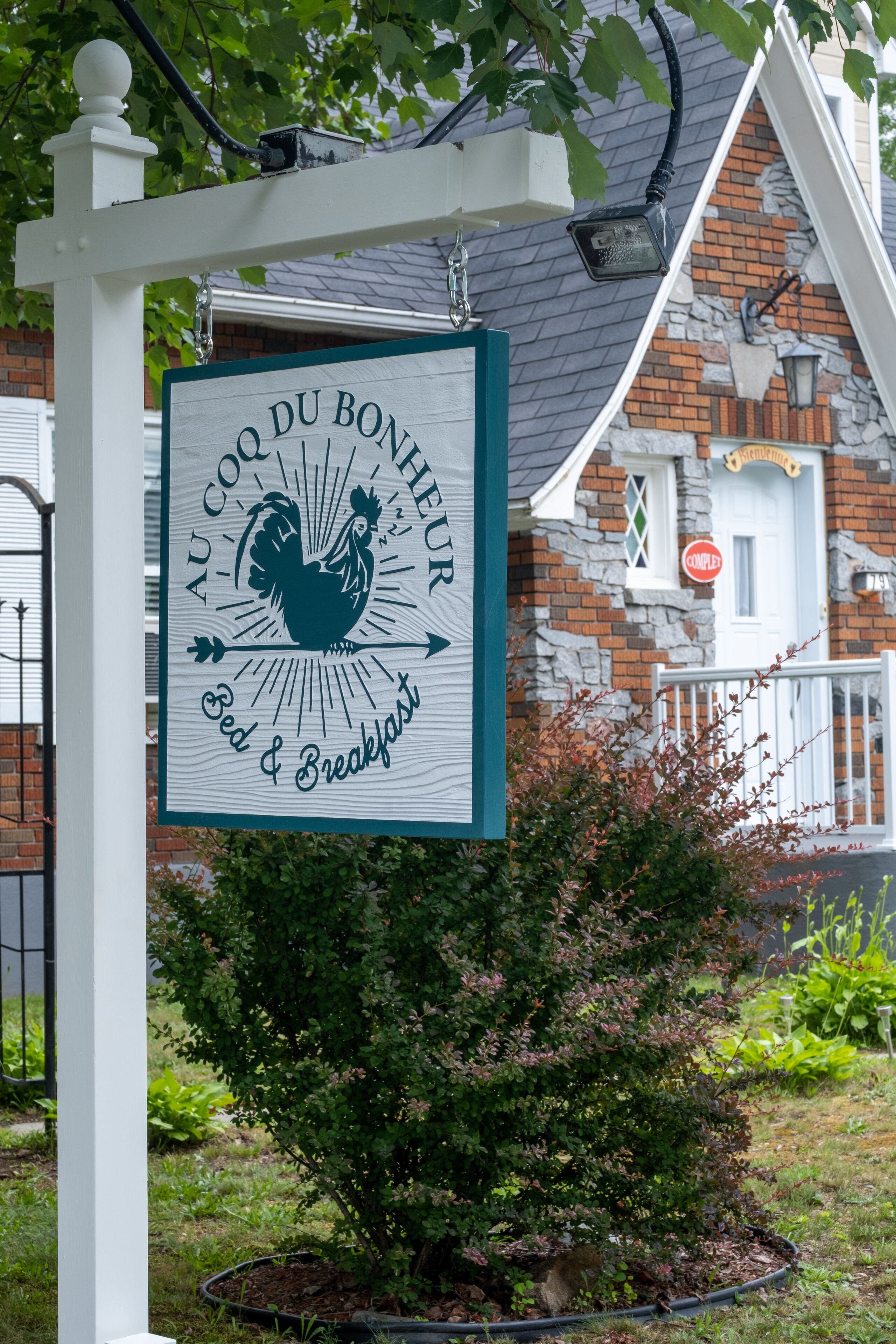 Une enseigne pour le « Au Coq du Bonheur Bed & Breakfast », avec un logo représentant un coq, placée sur un bâtiment en briques avec un porche blanc et de la verdure au premier plan.