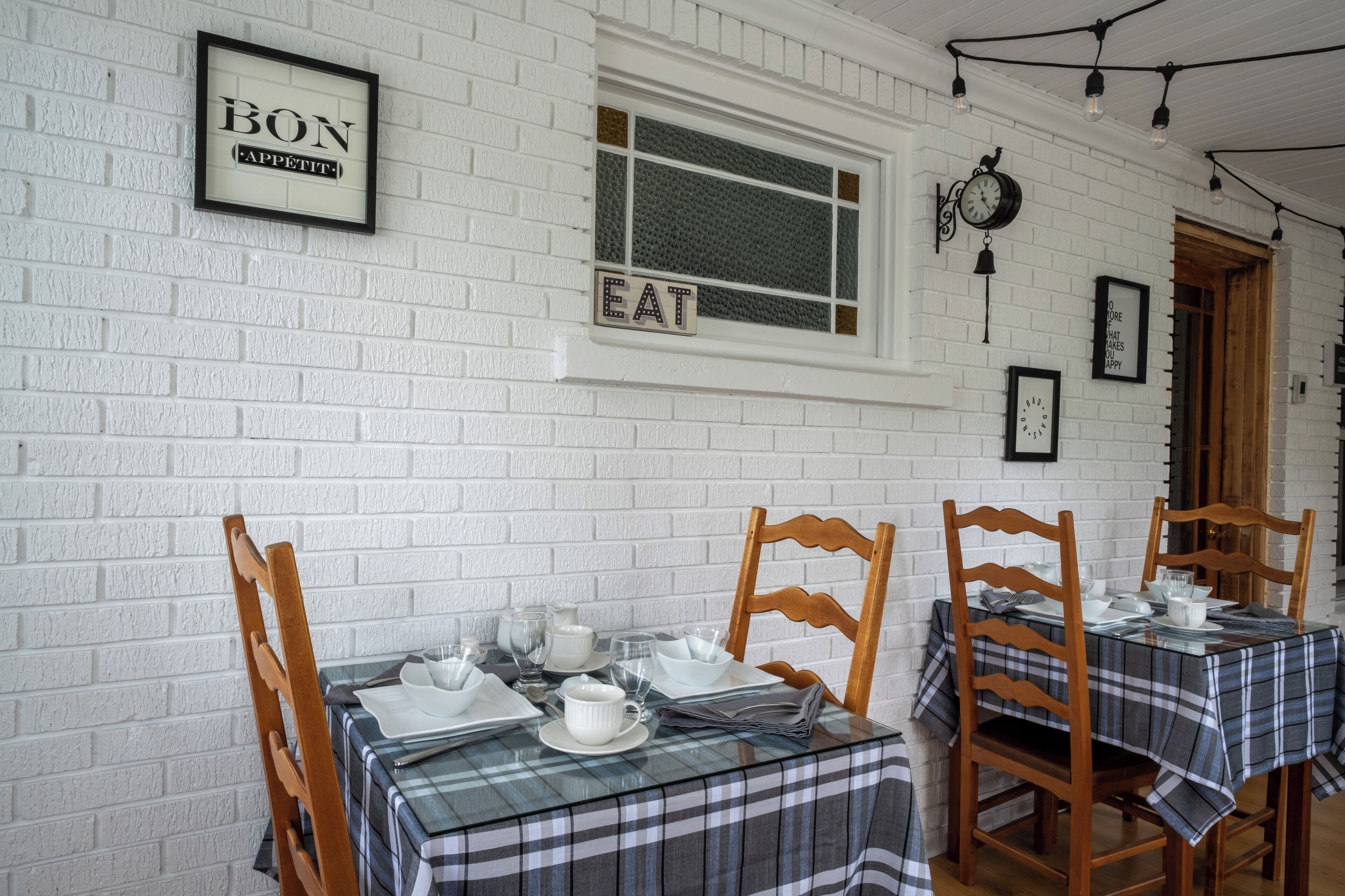A dining area with a white brick wall, framed signs reading "BON APPETIT" and "EAT", wooden chairs, and tables covered with plaid tablecloths.