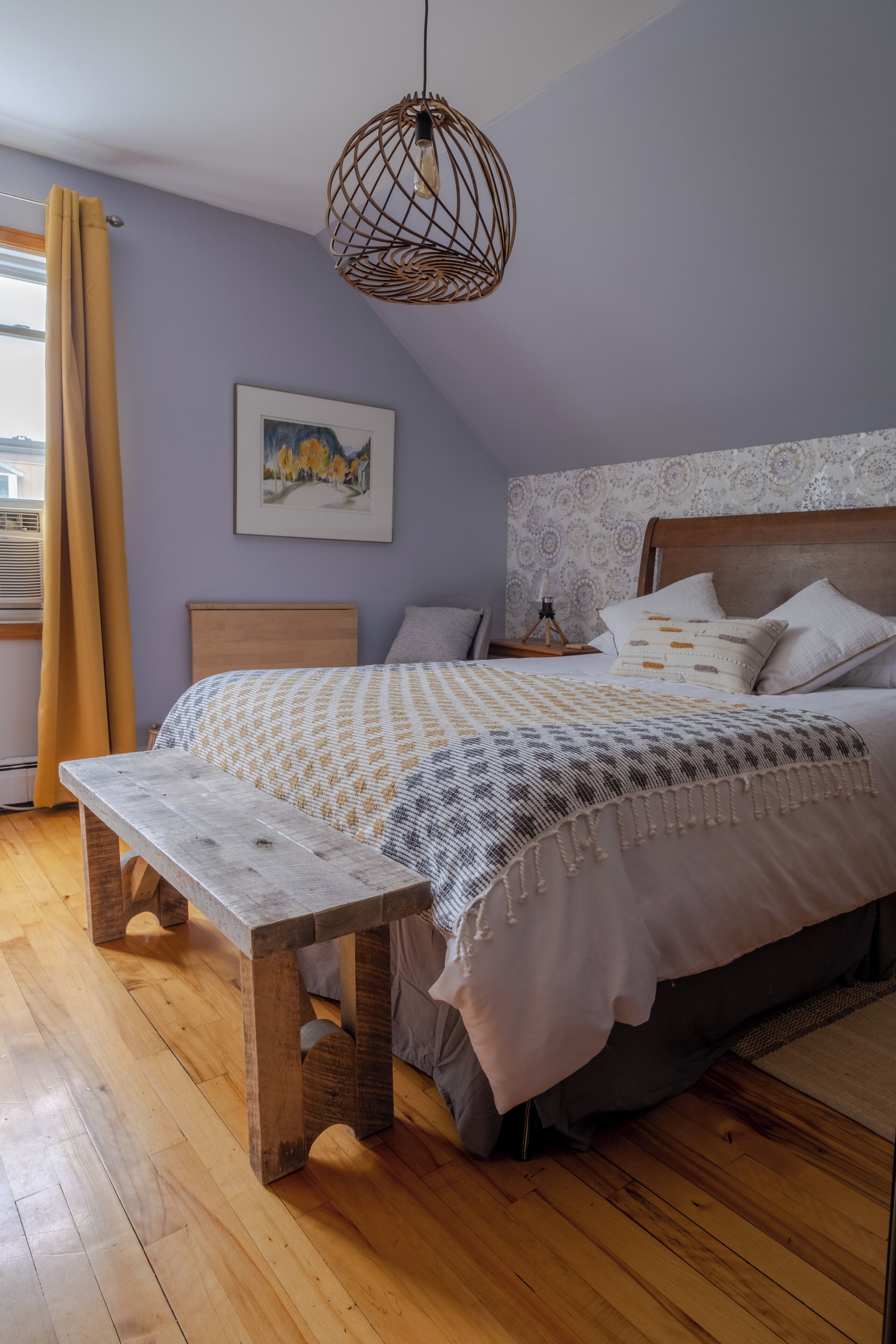 A bedroom with a wooden bench, a bed with patterned bedding, a hanging light fixture, and a framed picture on the wall.