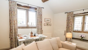 Un salon chaleureux avec un canapé beige, une table à manger dressée avec des assiettes et des tasses, des rideaux à motifs floraux et une fenêtre donnant sur un beau paysage.