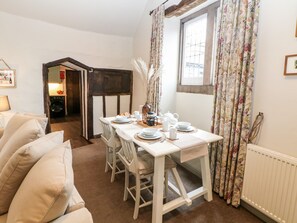 Un coin repas avec une table en bois dressée pour quatre personnes, des chaises blanches et une fenêtre ornée de rideaux à motifs floraux.
