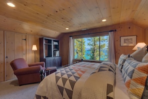 Un dormitorio acogedor con una cama grande, una silla, una estantería y un ventanal con vistas a árboles y agua.