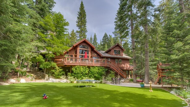 Une grande maison en bois dotée d’une terrasse et d’un escalier, entourée d’une végétation luxuriante et d’arbres.