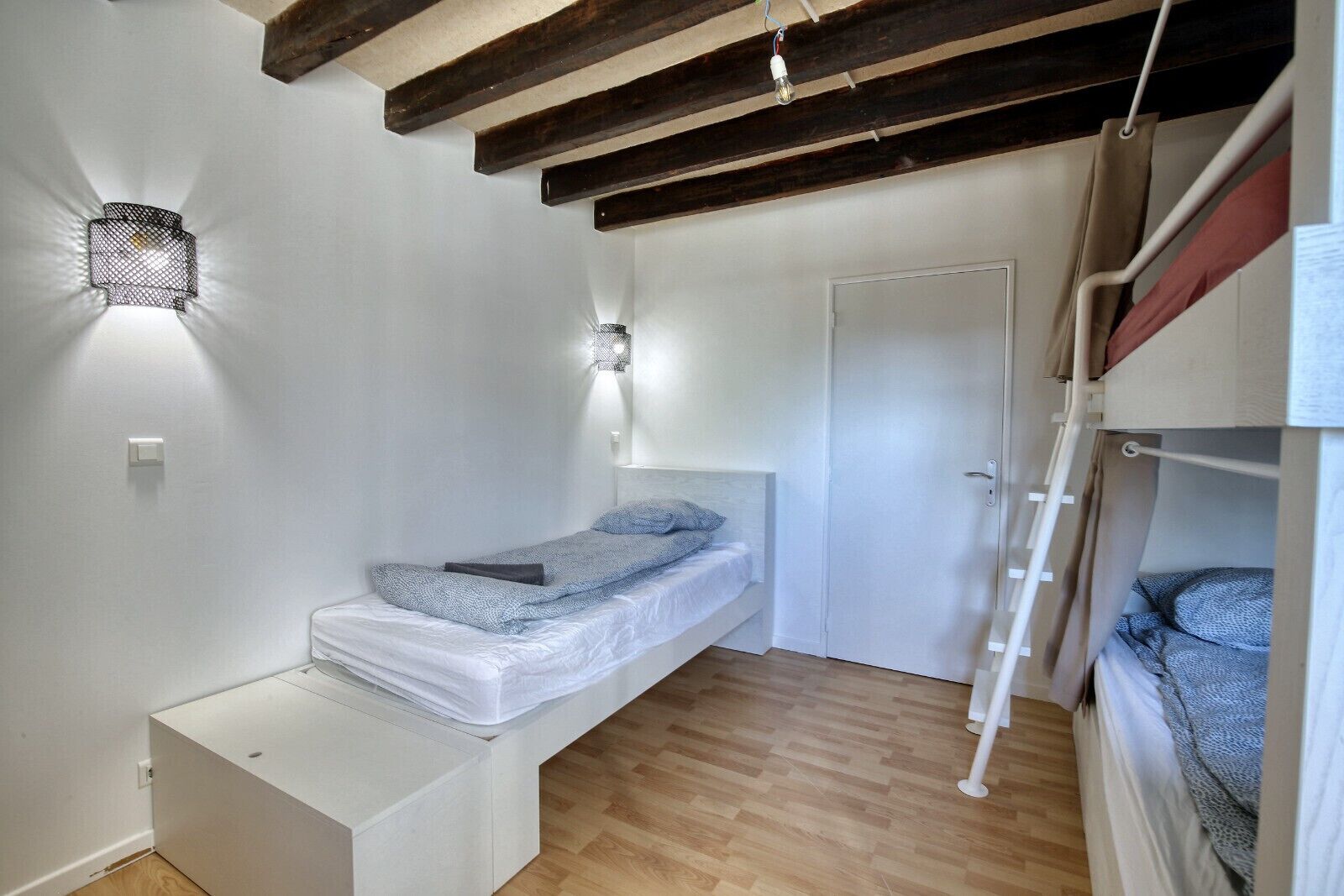 A compact bedroom with a bunk bed, a single bed, a small bedside table, a wall-mounted light, and a door.