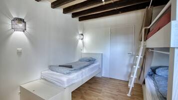 A compact bedroom with a bunk bed, a single bed, a small bedside table, a wall-mounted light, and a door.