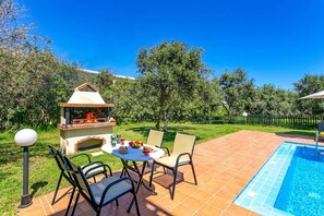 An outdoor patio with a barbecue grill, a round table with chairs, and a swimming pool. - Villa Ilos by Villa Plus (Pyrgos Psilonerou)