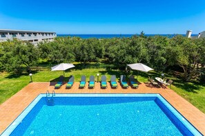 A rectangular swimming pool with a tiled edge, surrounded by a grassy area with lounge chairs and umbrellas. - Villa Ilos by Villa Plus (Pyrgos Psilonerou)