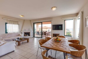 A living room with a sofa, a dining table with chairs, a fireplace, and a balcony with a view. - Villa Katerina Pente by Villa Plus (Kalives)